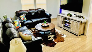 A living room with a black leather sofa, a round coffee table, a flat-screen TV, and a wooden cabinet with books. - Sports Themed 3-Bedroom Home in Arlington near AT & T Stadium & Other Sports  (Arlington)