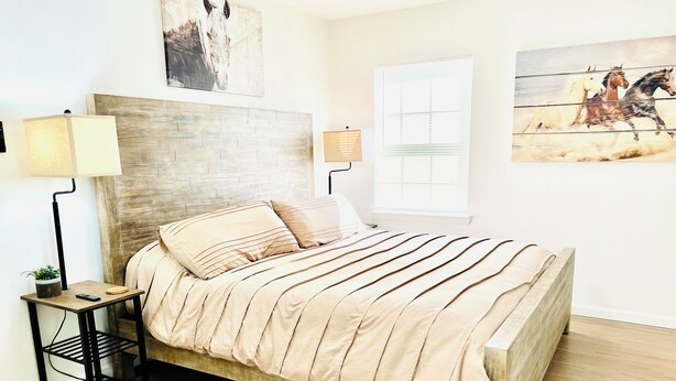 A bedroom with a wooden headboard, a bed with striped bedding, a bedside table with a lamp, and artwork on the walls. - Sports Themed 3-Bedroom Home in Arlington near AT & T Stadium & Other Sports  (Arlington)