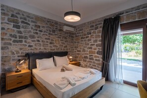 A bedroom with a stone wall, a bed with white linens, a bedside lamp, and a view through a window with curtains. - Villa Liatiko With Private Pool (Agios Vasileios)