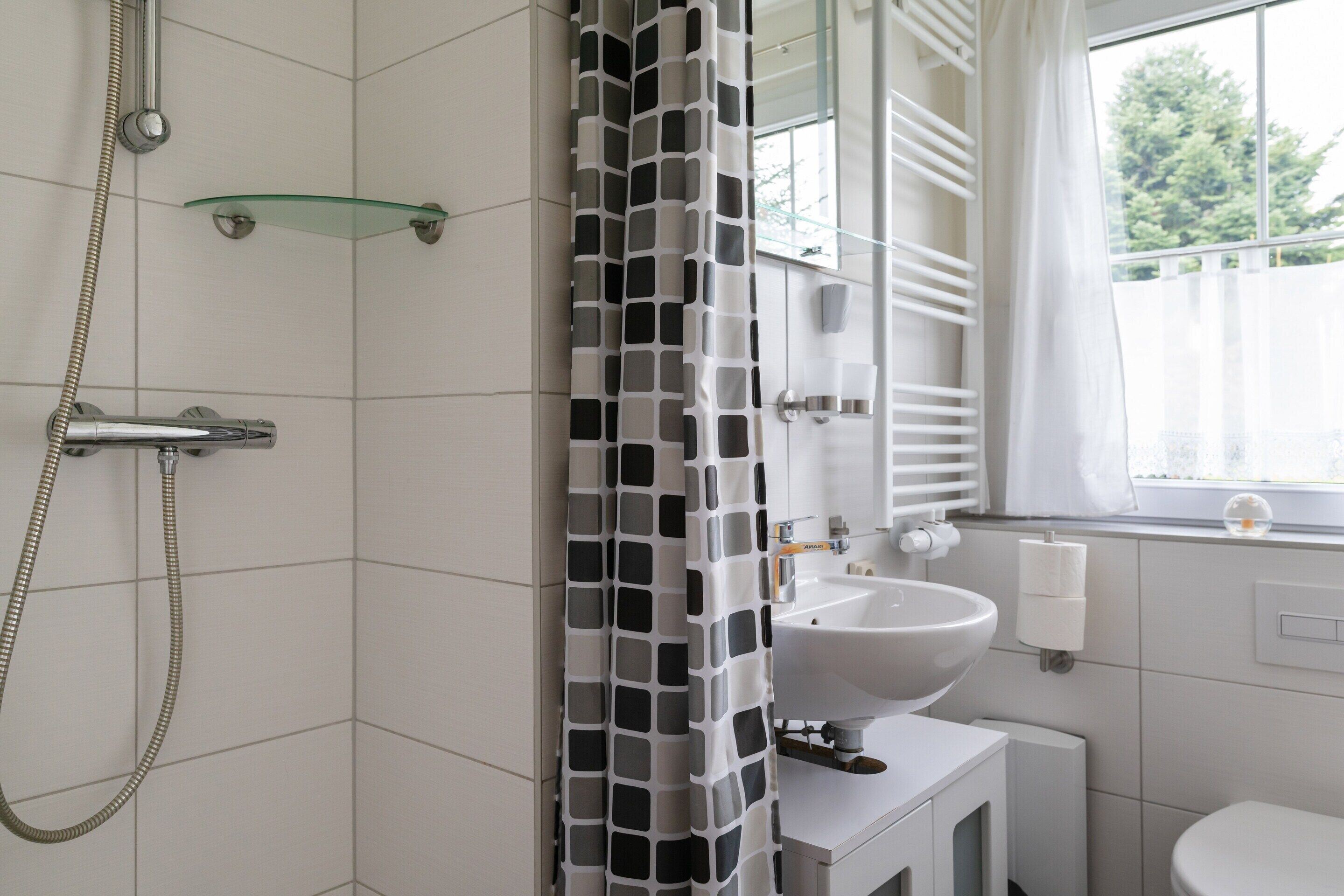 Bagno con doccia, ripiano in vetro, lavandino e finestra vista alberi.