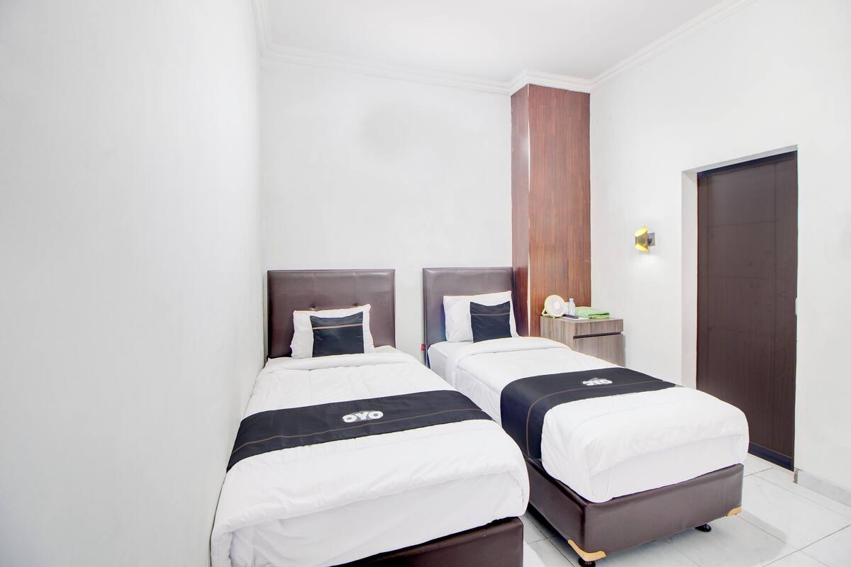 Habitación de hotel con dos camas, un armario de madera, una mesita de noche con lámpara y una puerta.