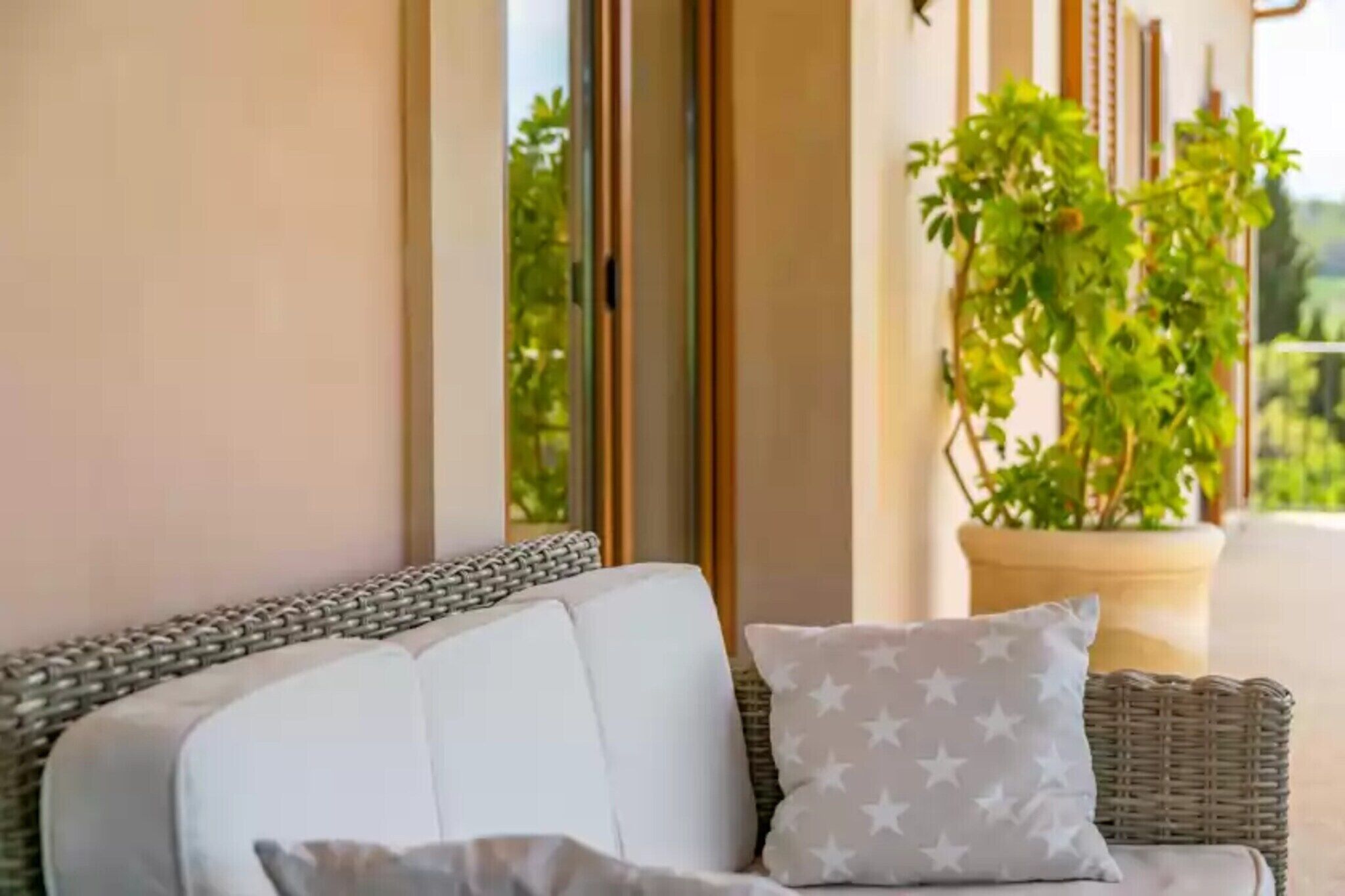 A wicker sofa with white cushions and a star-patterned pillow, positioned near a large potted plant on a balcony with a view of greenery.