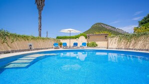 A clear blue swimming pool with three blue lounge chairs and a white umbrella, surrounded by a low stone wall and greenery, with a mountain in the background.