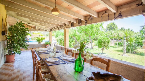 An outdoor dining area with a wooden table set for two, surrounded by chairs, with a view of a garden and a pool in the background.
