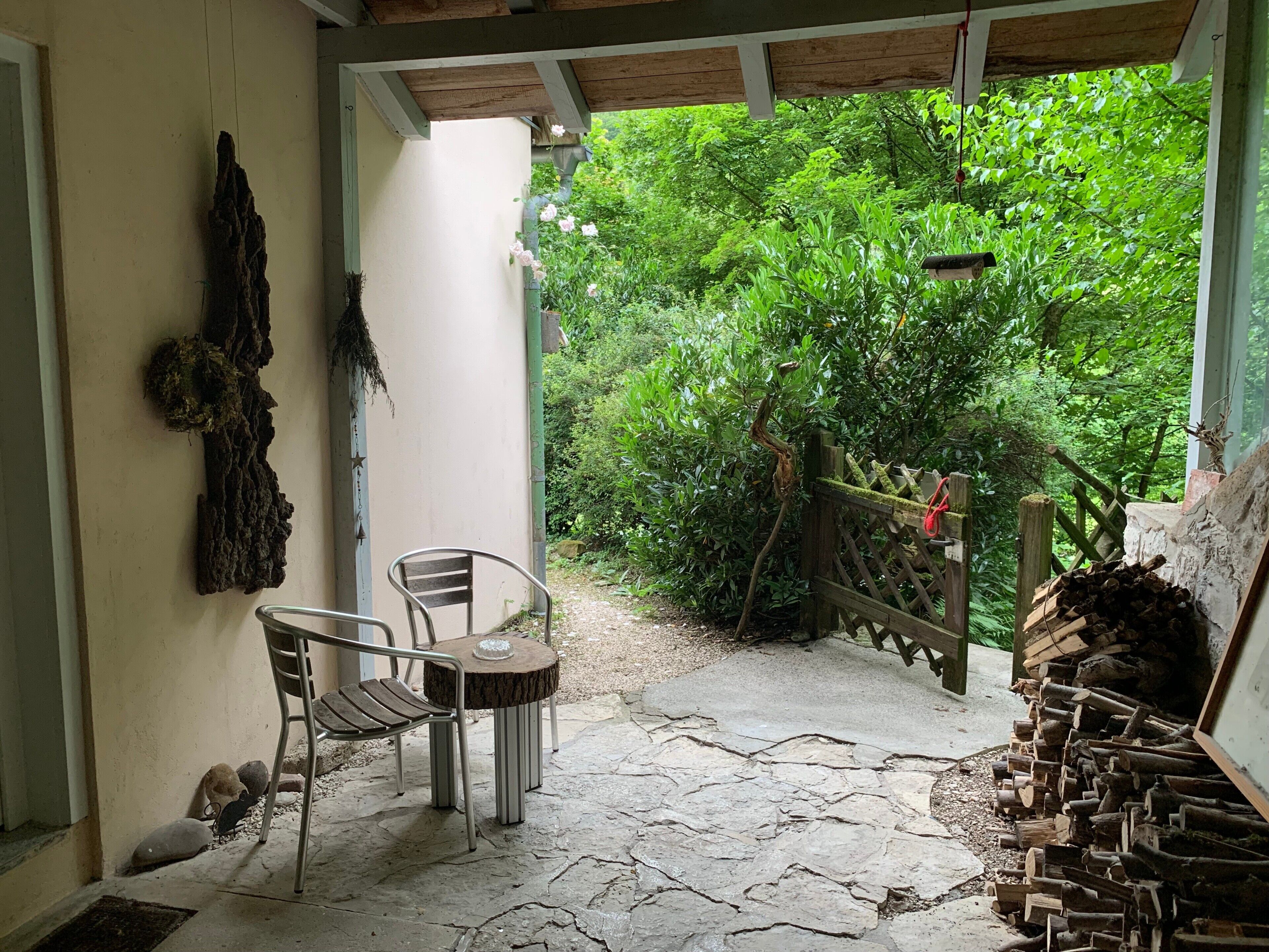 A covered patio with a stone floor, two metal chairs, a wooden table, a rustic wooden fence, and a pile of firewood.