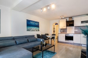 Una sala de estar moderna con un sofá gris, una mesa de comedor con sillas, una cocina con armarios blancos y un cuadro en la pared.