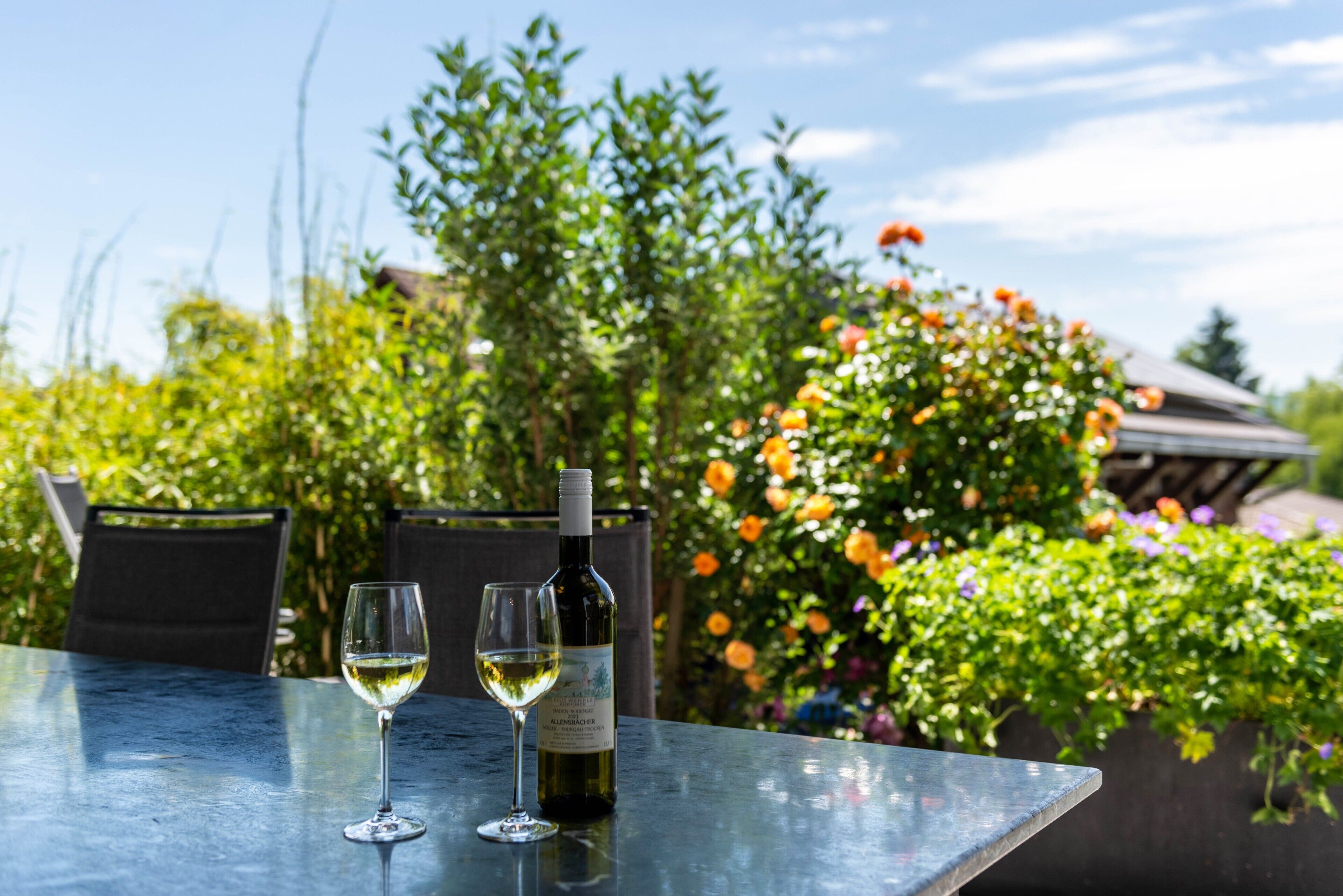 A table with two wine glasses and a bottle, surrounded by greenery and flowers.