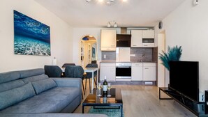 Uma sala de estar moderna com um sofá, uma mesa de centro com copos de vinho, uma televisão de tela plana e uma cozinha ao fundo.