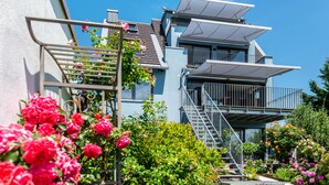 Ein modernes, mehrgeschossiges Gebäude mit einer Treppe, die zu einem Balkon führt, umgeben von üppiger Vegetation und blühenden Pflanzen.