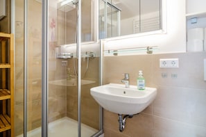 Un bany modern amb un compartiment de dutxa de vidre, un lavabo blanc amb dispensador de sabó i un mirall sobre el lavabo.