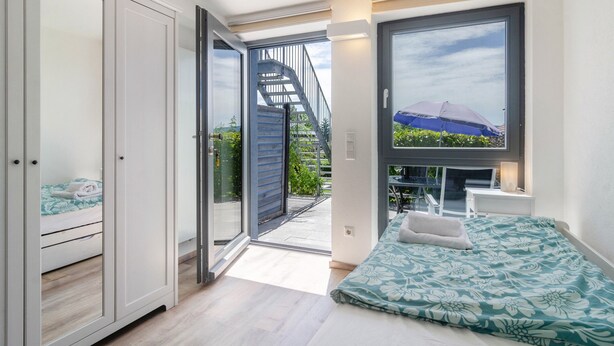 Ein Schlafzimmer mit Bett, Kleiderschrank, Schreibtisch und einem Fenster mit Blick auf eine Treppe und Grünflächen.