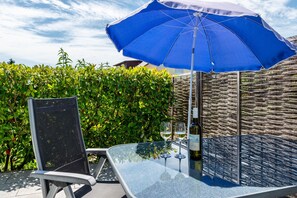 A glass table with two wine glasses and a bottle, a blue umbrella, and a chair in a sunny outdoor setting. - Studio Apartment with Private Terrace and Wi-Fi (Allensbach)
