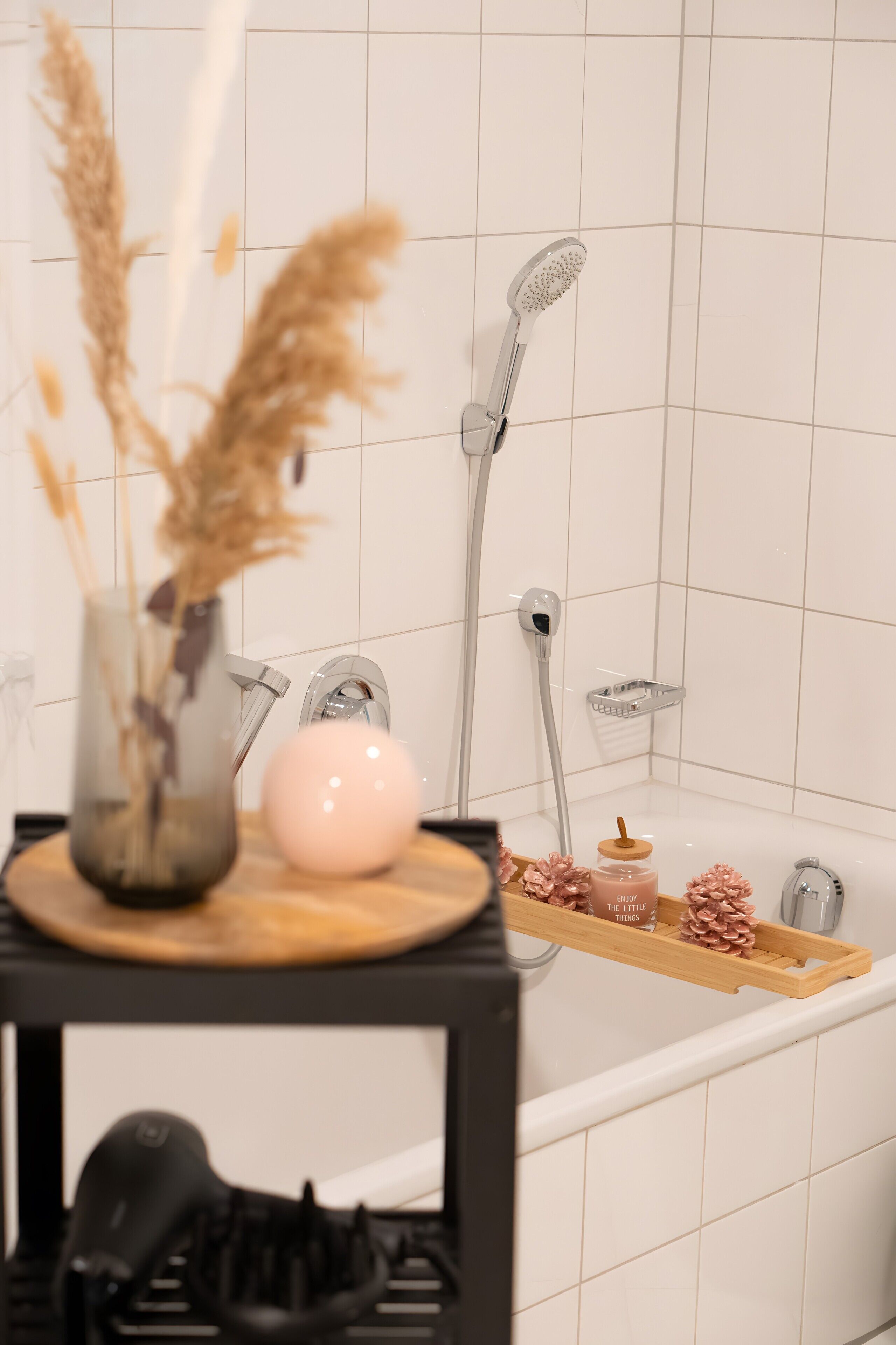 Une salle de bain avec une douche, un lavabo, un vase contenant des plantes séchées, un distributeur de savon rose et un plateau avec des pommes de pin et une bouteille.