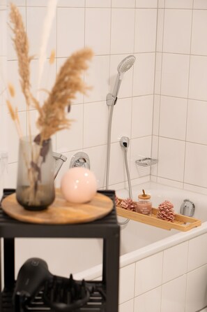A bathroom with a shower, a sink, a vase with dried plants, a pink soap dispenser, and a tray with pine cones and a bottle. - Apartment 'Wohnung Im Rheintal' with Balcony and Wi-Fi (Küssaberg)