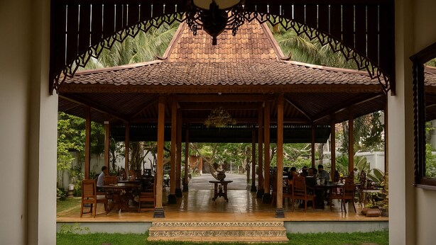 A covered outdoor seating area with wooden tables and chairs, a tiled roof, and a view of a garden. - The Mangkoro Hotel by Sinergi (Banguntapan)