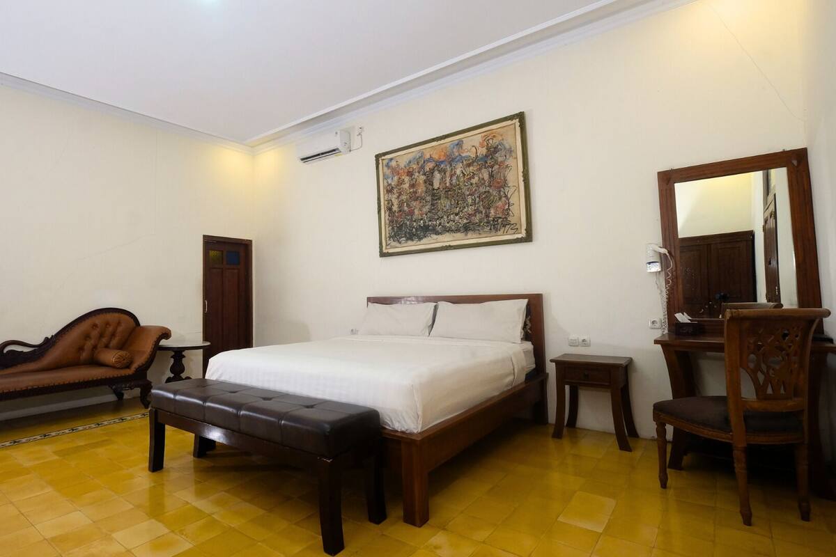 A hotel room with a bed, a wooden bench, a desk with a chair, and a painting on the wall.