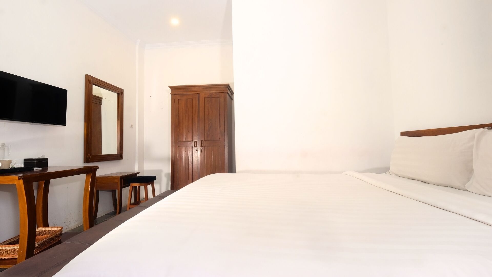 A hotel room with a large bed, a wooden desk with a chair, a flat-screen TV mounted on the wall, and a wooden wardrobe.