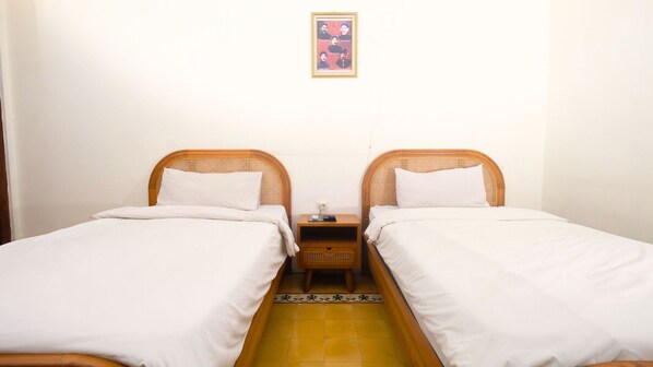 A room with two single beds, each with white bedding and wooden headboards. There is a small wooden nightstand between the beds with a lamp and a book. The floor is tiled in a yellow pattern. - The Mangkoro Hotel by Sinergi (Banguntapan)