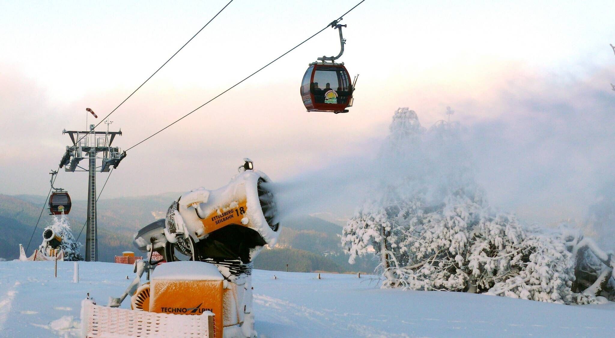 Um teleférico com um funicular ao fundo, uma árvore coberta de neve e um preparador de neve em primeiro plano.