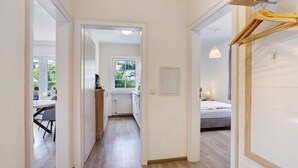 Un couloir d’hôtel avec un plancher en bois, donnant sur une salle à manger, une cuisine et une chambre à coucher avec un lit et un luminaire suspendu.