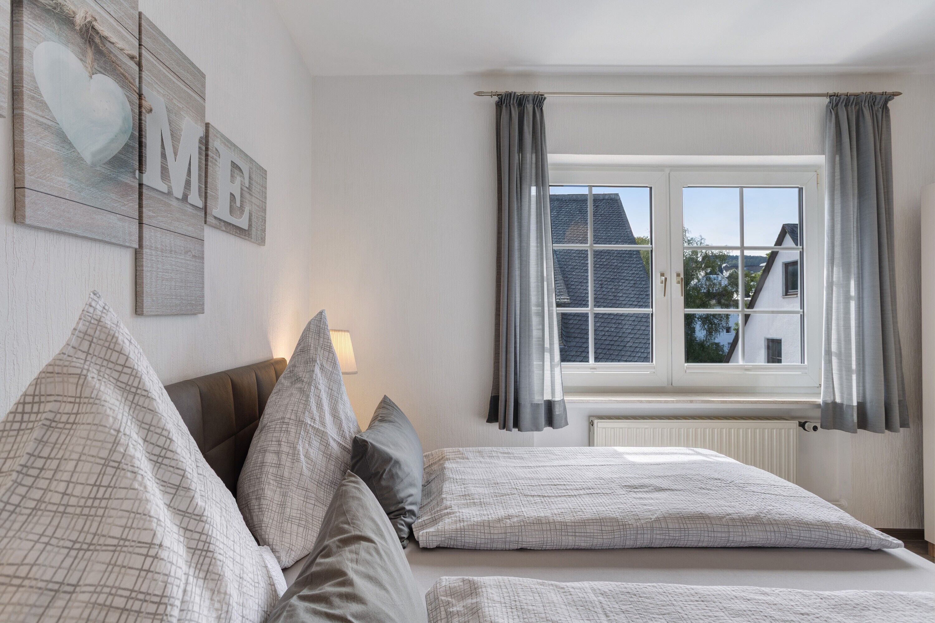 A bedroom with two beds, a window with curtains, a radiator, and a wall with a heart-shaped decoration.