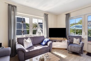 Un salon avec un canapé gris, un fauteuil plus petit, un téléviseur à écran plat et de grandes fenêtres avec des rideaux.