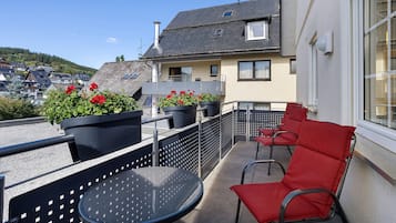 Un balcon avec une table ronde et des chaises rouges, des plantes en pot et une vue sur un quartier résidentiel avec des maisons et une colline.