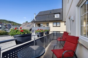 A balcony with a round table and red chairs, potted plants, and a view of a residential area with houses and a hill.