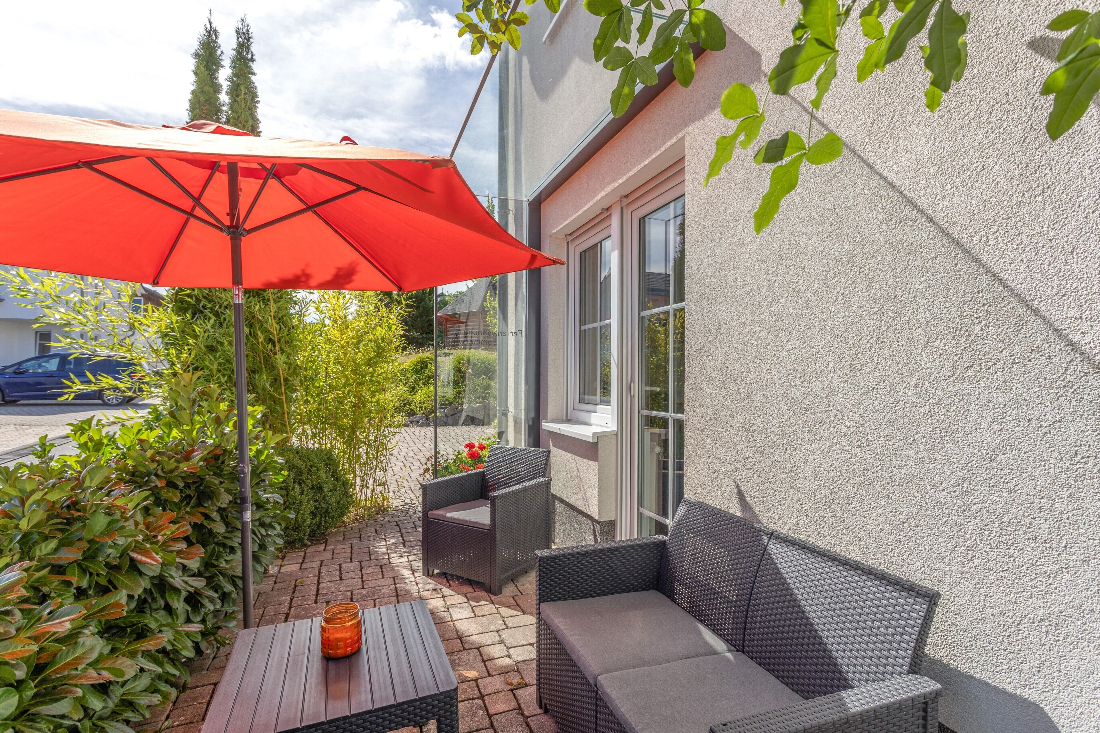 Et udendørs siddeområde med en rød parasol, fletningsmøbler og udsigt til gaden.