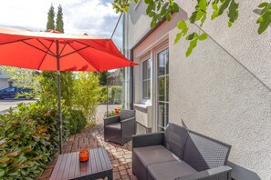 Un espace extérieur aménagé avec des sièges, un parasol rouge, des meubles en osier et une vue sur la rue.