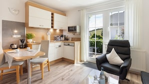 Une cuisine moderne avec des meubles blancs, un coin repas comprenant une table et des chaises, et un salon avec un fauteuil noir et une table basse en verre.