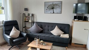 Un salon avec un canapé gris foncé, deux fauteuils noirs, une table basse sur laquelle se trouvent une bouteille, des verres et un petit objet décoratif. Un tableau encadré représentant un arbre est accroché au mur, et un téléviseur est fixé au-dessus d’un meuble.