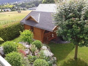 A wooden cabin with a sloped roof, surrounded by a well-maintained garden with various plants and a large tree.