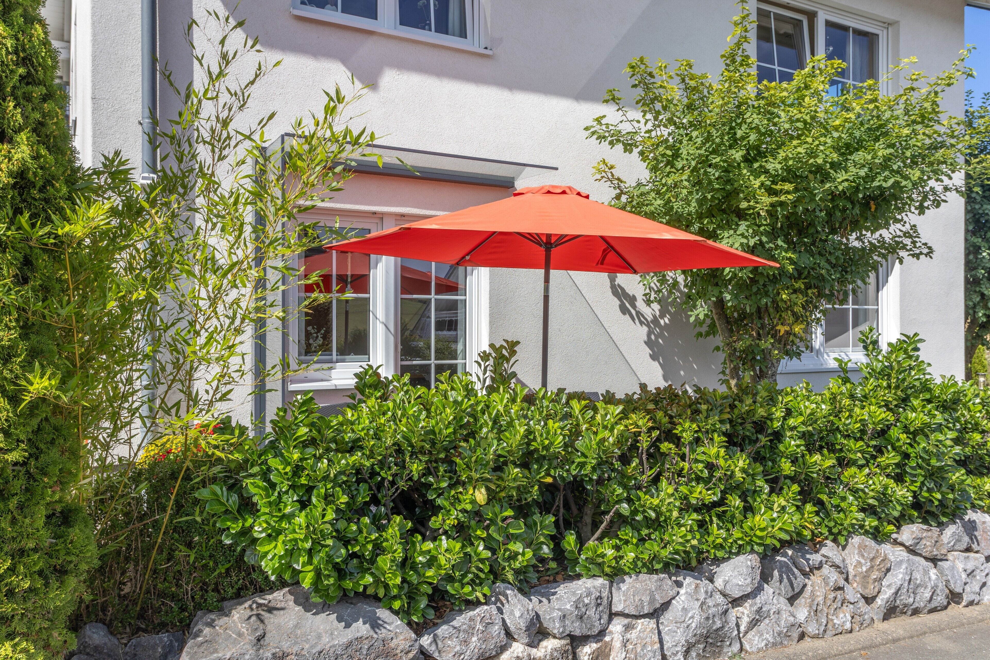 A building with a stone wall, a red umbrella, and green foliage.