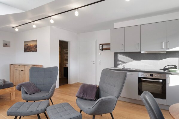 A modern kitchen with grey cabinets, a built-in oven, and a dining area with grey upholstered chairs.