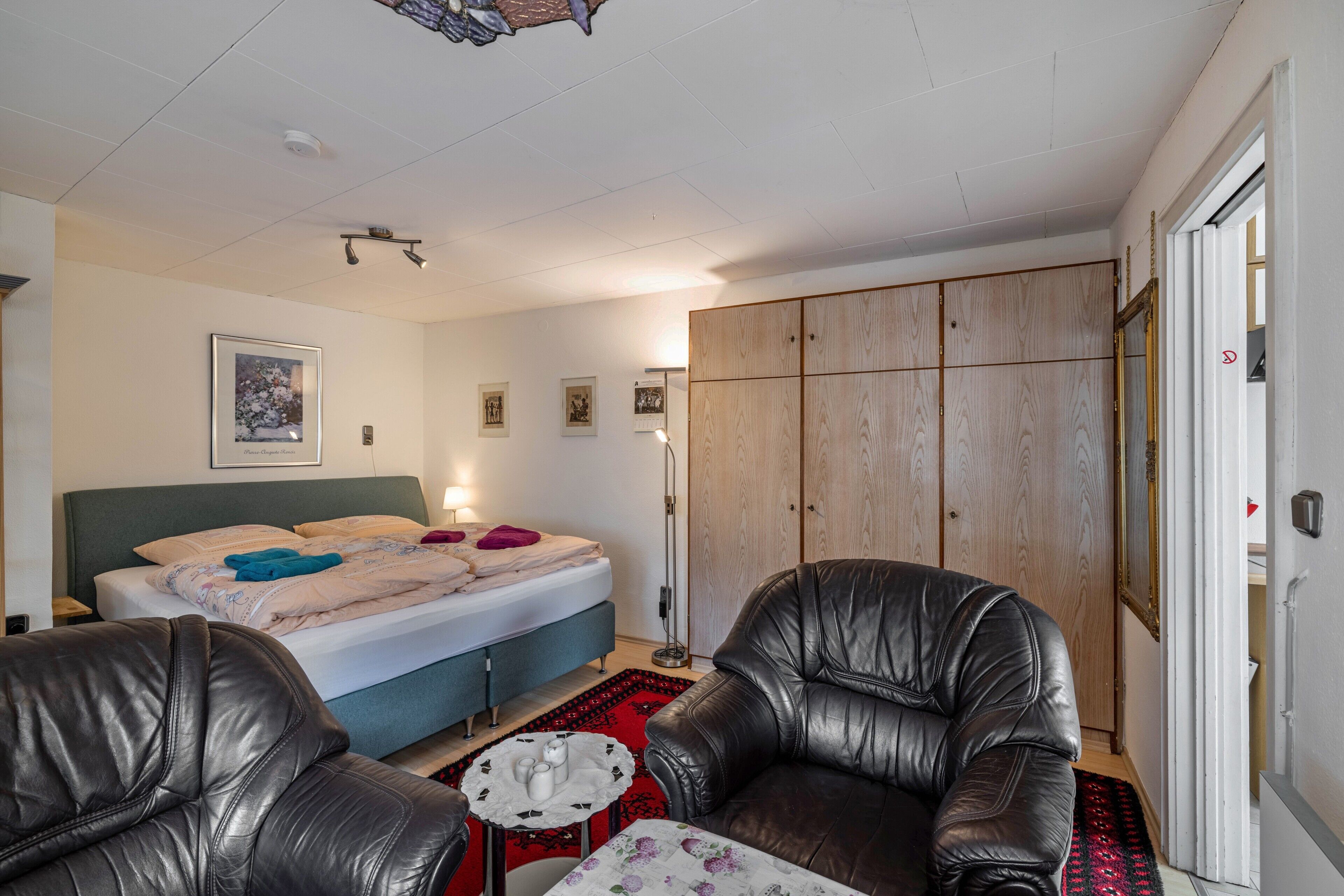 Une chambre à coucher comprenant un lit, deux fauteuils en cuir, une table de chevet, un tapis rouge et une armoire.