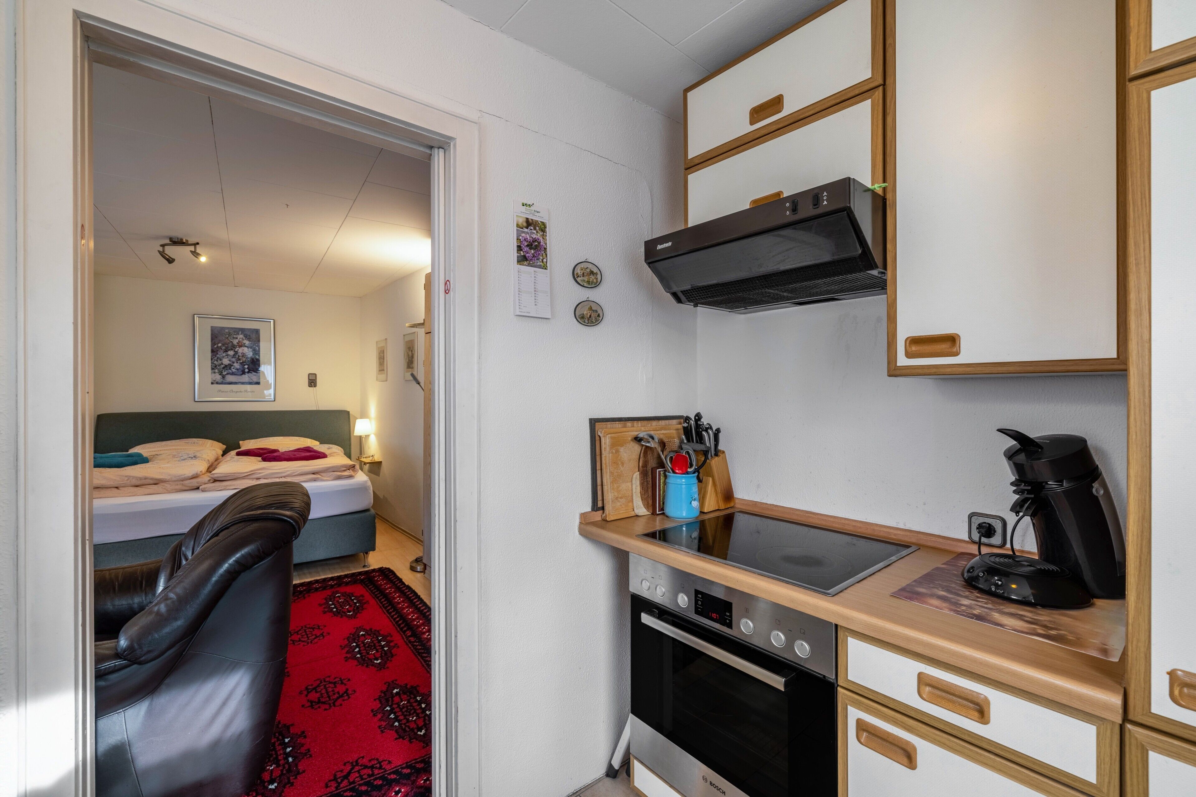 A compact hotel room with a kitchenette, a bed, a red patterned rug, and a black leather chair.