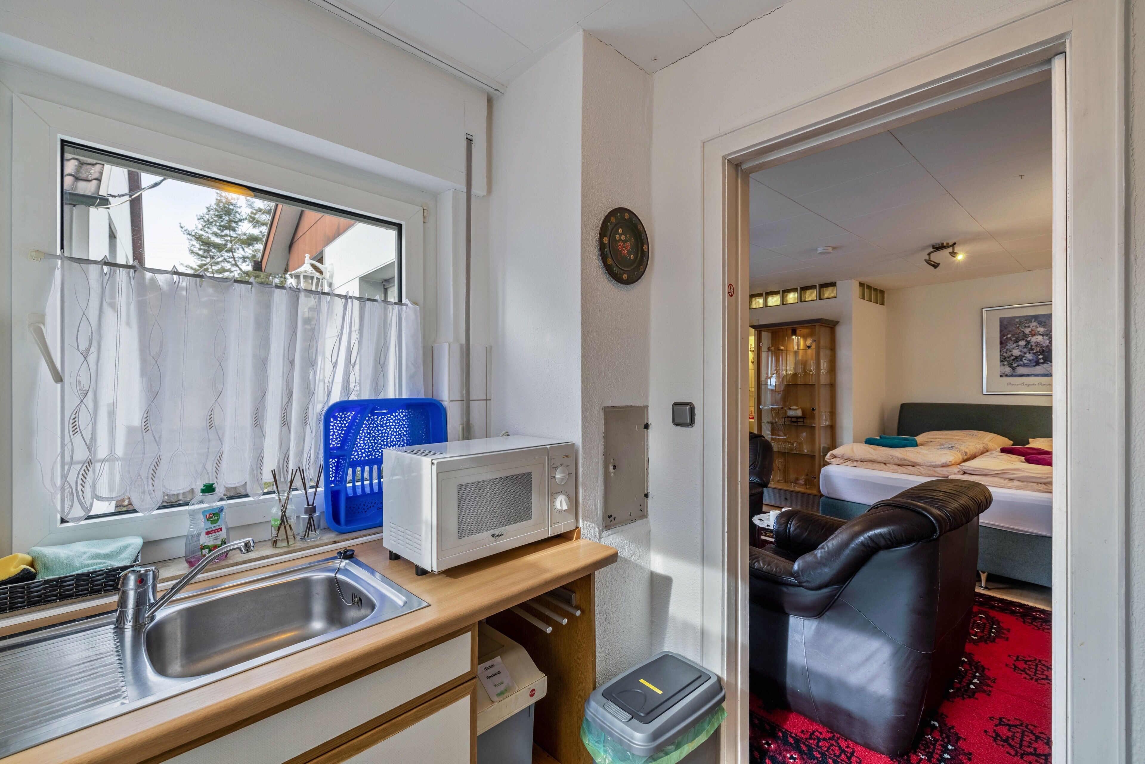 A compact kitchenette with a microwave, sink, and window with curtains.