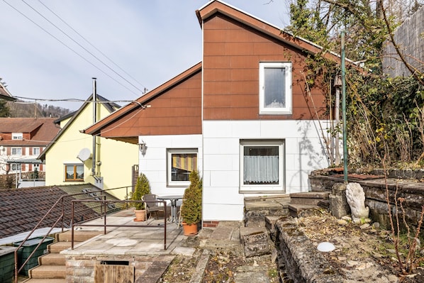 A two-story house with a brown roof, white walls, and a small garden area with steps leading to the entrance.