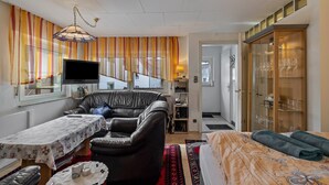 Een kamer met een leren bank, een klein tafeltje met een tafelkleed met bloemenprint, een bed met een bedspreide met dessin, een televisie en een kast met glazen deuren waar glazen in staan.