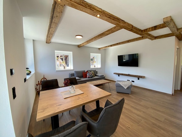 A modern living room with exposed wooden beams, a large flat-screen TV mounted on the wall, a grey sofa, and a wooden dining table with chairs.