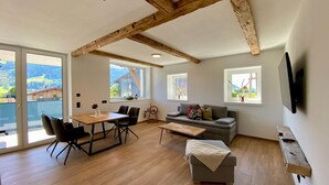 A spacious living room with a wooden ceiling, a gray sofa, a wooden dining table with chairs, and large windows offering a view of the outdoors.