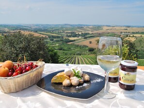 A table with a basket of cherries, a plate of pastries, a glass of white wine, and two jars of jam with a scenic vineyard view in the background.