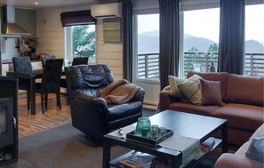 Una sala de estar moderna con un sofá de cuero, un sofá marrón y una mesa de centro. Hay un comedor con una mesa y sillas, y una cocina con armarios y estufa. Grandes ventanas ofrecen vistas al exterior.