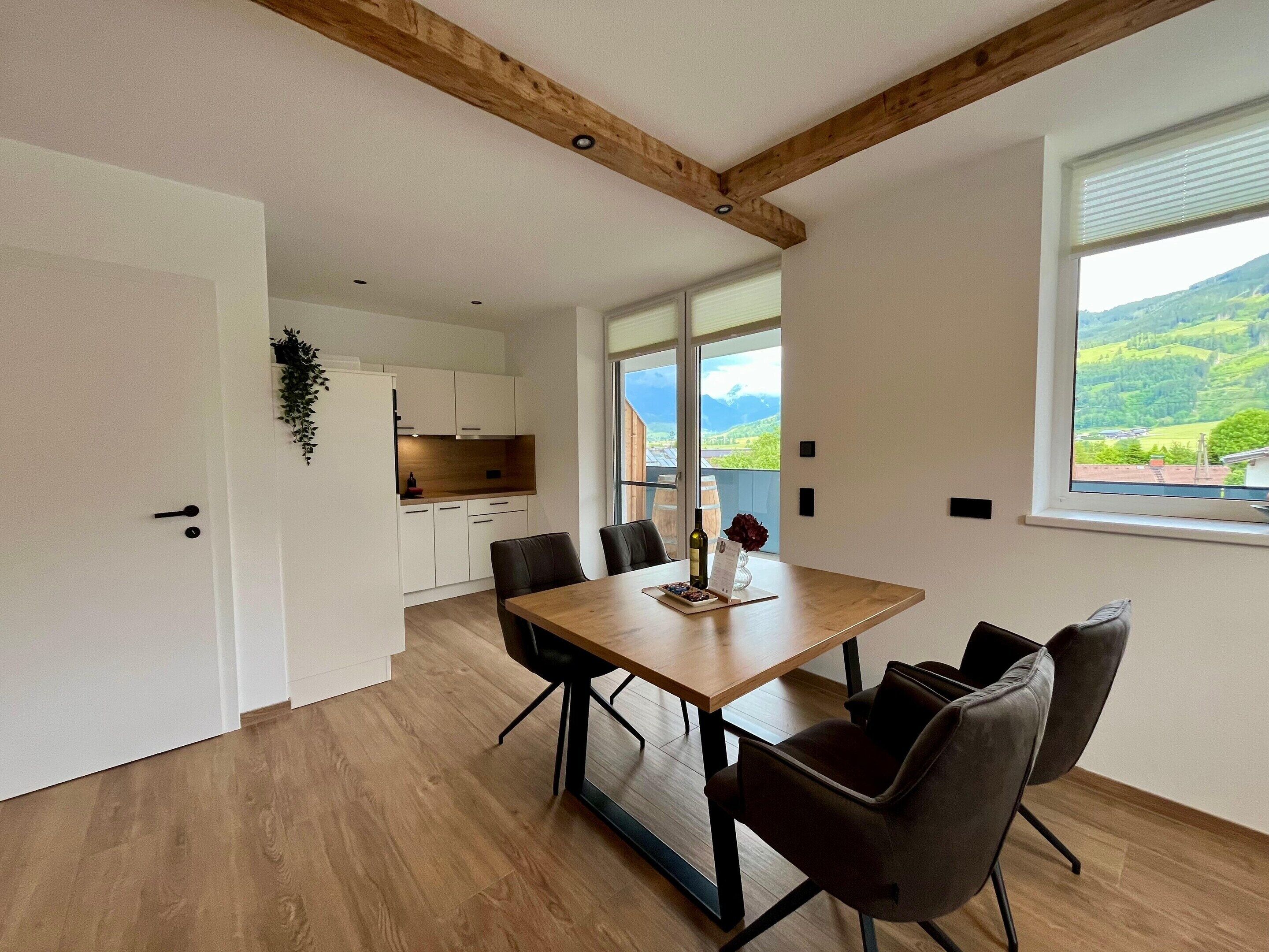Uma área de jantar moderna com mesa e cadeiras de madeira, vista para o campo através de janelas amplas e uma cozinha com armários brancos.