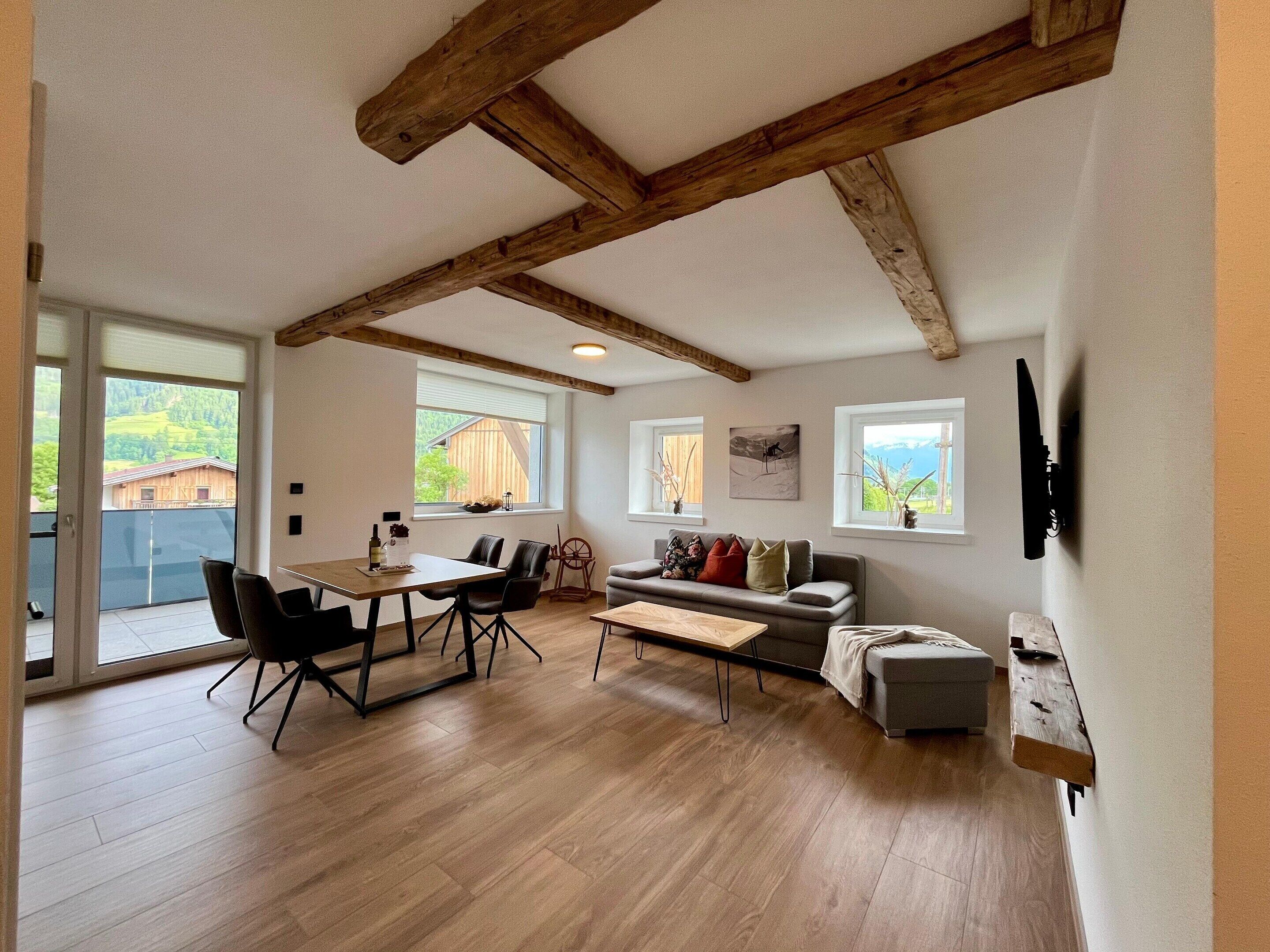 Un salón amplio con techo de madera, una mesa de comedor con sillas, un sofá y un televisor fijado a la pared.
