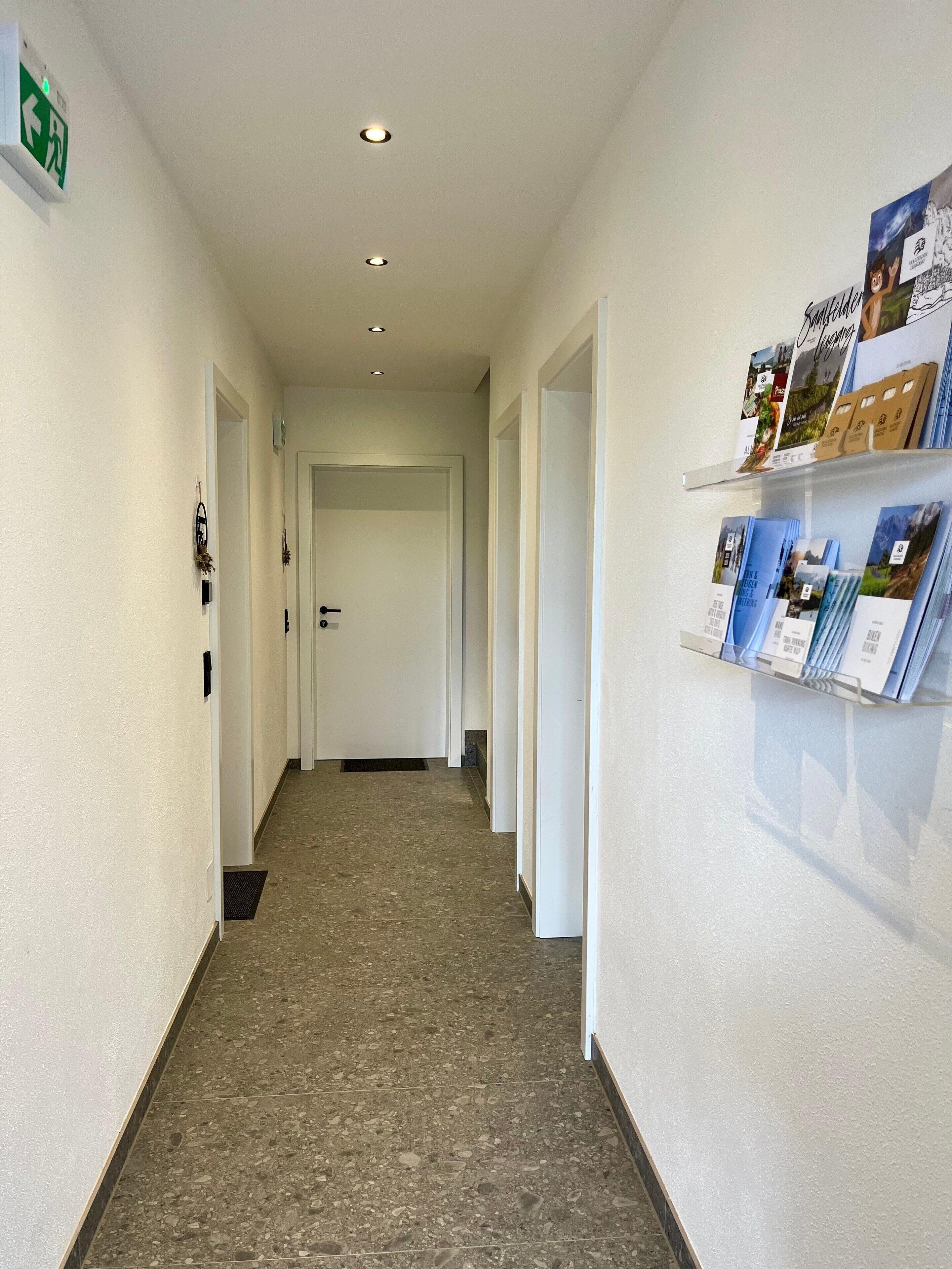 Un corridoio con pavimento piastrellato, una porta in fondo e una bacheca con brochure sulla parete destra.