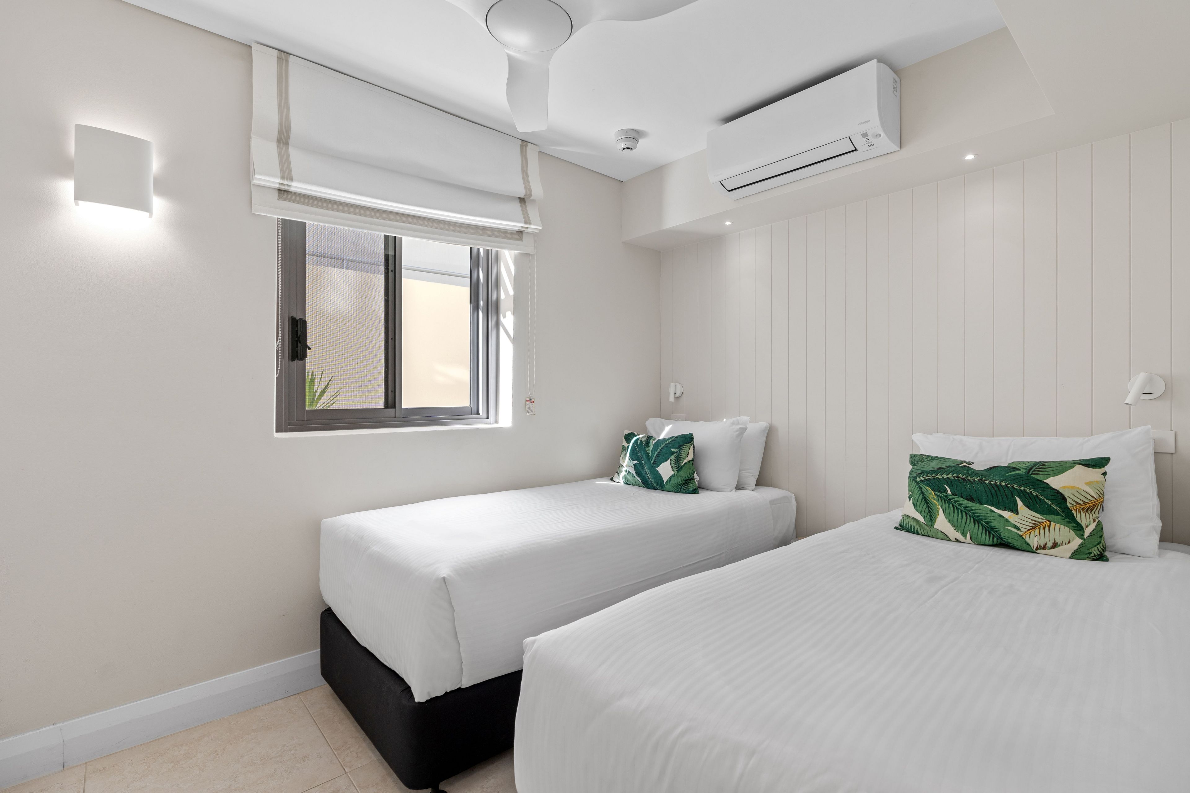 Een slaapkamer met twee bedden, een raam met zonwering, een airconditioningapparaat en een plafondlamp.