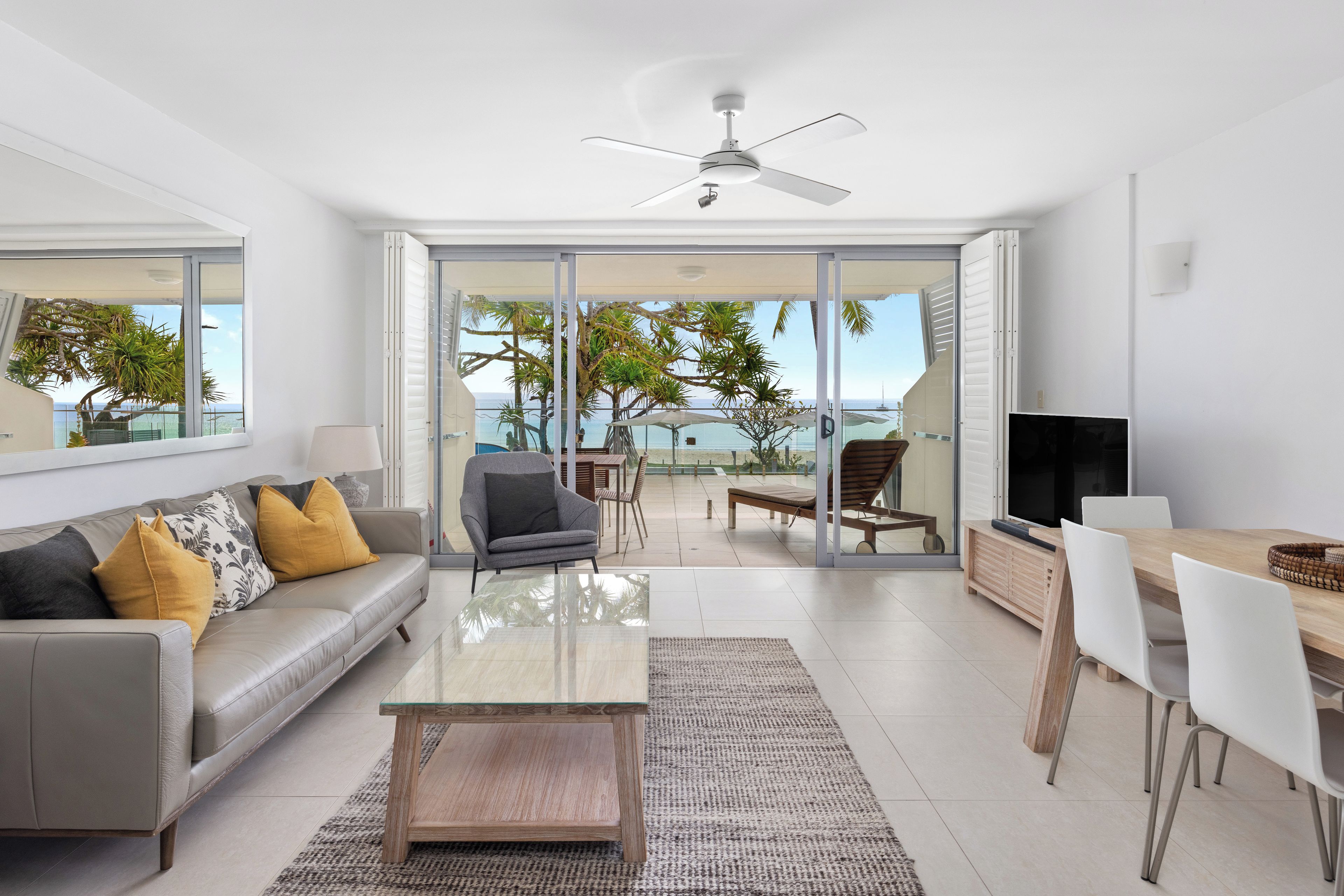A modern living room with a sofa, coffee table, and a TV. There is a balcony with a view of the sea and palm trees.