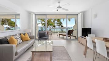 A modern living room with a sofa, coffee table, and a TV. There is a balcony with a view of the sea and palm trees.